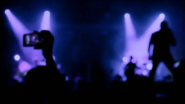 Ve akıllı telefon kayıtlarıyla canlı müzik performansı. Sahnede mor ışıklarla aydınlatılmış bir rock şarkıcısının bulanık silueti.