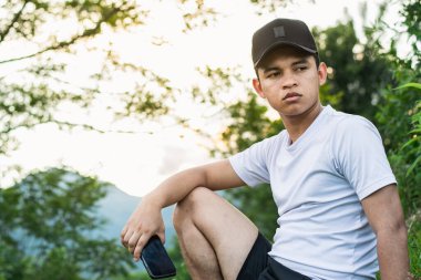 Genç Latin bir adam dağın tepesinde oturmuş arka planda altın bir günbatımı olan güzel doğayı seyrediyor. Elinde cep telefonu olan bir çocuk. siyah başlık ve beyaz tişört.