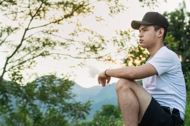 Genç Latin erkek, üniversite öğrencisi, tropikal ormanın ortasında oturuyor, arka planda günbatımı, gururla Kolombiya dağlarının güzel doğasına bakıyor..
