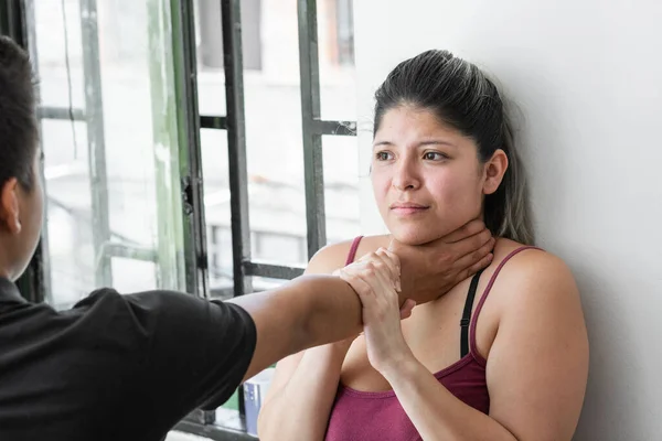 latina woman with a scared face, while her husband grabs her by the neck. girl trying to defend herself from her aggressive boyfriend, trying to grab his hand to get loose.