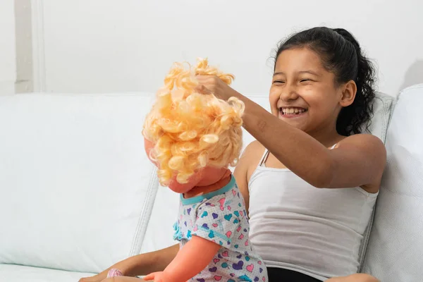 大きな笑顔でソファで人形と遊んでいるラティーナの女の子 ブルネット来る彼女のおもちゃ ロイヤリティフリーのストック画像