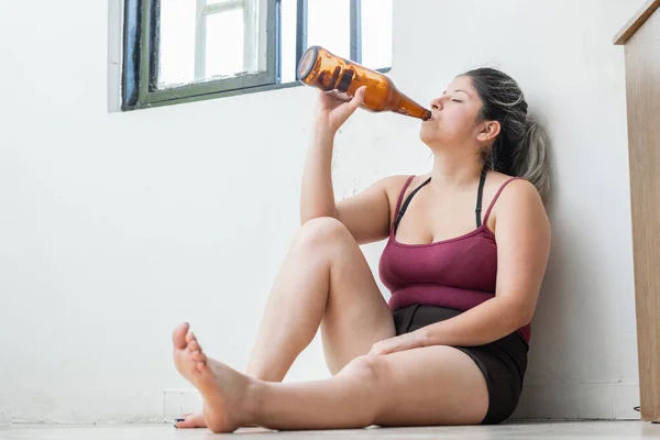 woman sitting on the floor drinking from a beer bottle, depressed girl venting her pain in alcohol. latina woman sad about losing a loved one.