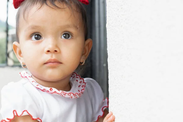 dark-haired baby, with curious look. girl looking distrustful. standing on the side of a wall.
