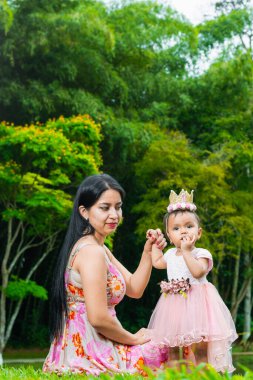 Latin anne çimlerin üzerinde oturmuş gururla kumral bebeğine bakıyor, büyük ağaçlarla yeşil bir parkta geziniyor..