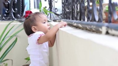 Kahverengi tenli, etrafı keşfeden ve etrafındaki nesnelere şaşıran güzel Latin bir kız bebek. Küçük kız evinin balkonunda tek başına duruyor..