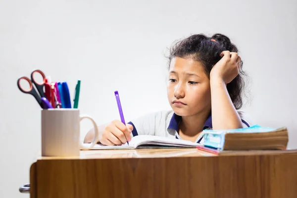latina brunette student, very bored and sad doing her homework. little girl at her wooden desk writing on her notebook.