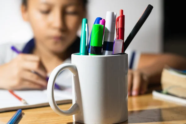 detailed view of a white cup full of pens and school supplies, in the background a little Latina student girl writing on her notebook.