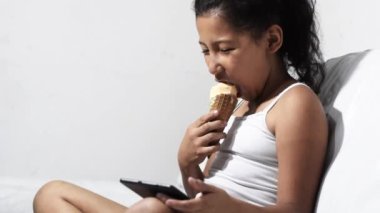 happy, smiling brunette Latina girl eating an ice cream cone while looking at her tablet, sitting on the sofa at home.