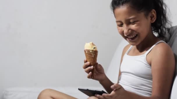 美しいブルネットのラティーナの女の子家でソファに座ってアイスクリームを食べながら彼女のソーシャルネットワークをタブレットで見て — ストック動画