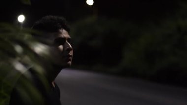 close-up of a pensive teenager, sitting on the side of the road, as cars pass by him on a quiet night.