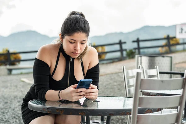 young latina woman, looking at cell phone, leaning on the table while waiting for her order alone