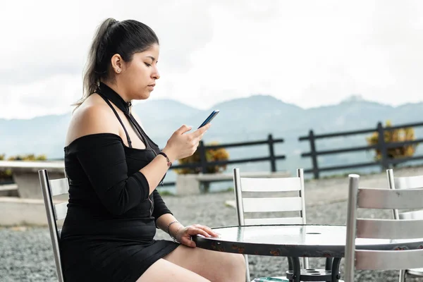 young latina woman looking at her cell phone while sitting in an outdoor caf