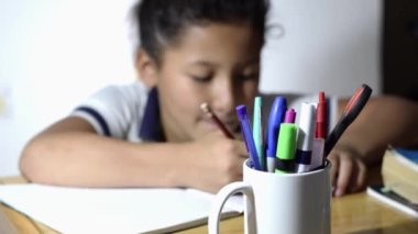 close-up of a cup full of colored pencils, in the background a little latin girl studying on a wooden desk