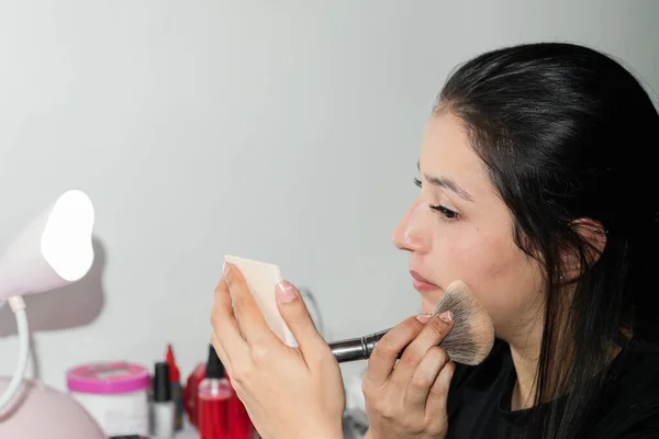 young latina woman applying a contour with a brush while looking at herself in a small mirror