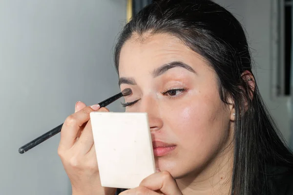 face of a latin woman applying makeup to her eyelids with a small brush while looking in the mirror
