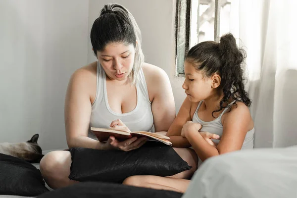 Chica Morena Mirando Curiosamente Mientras Madre Lee Libro Sentada Cama — Foto de Stock