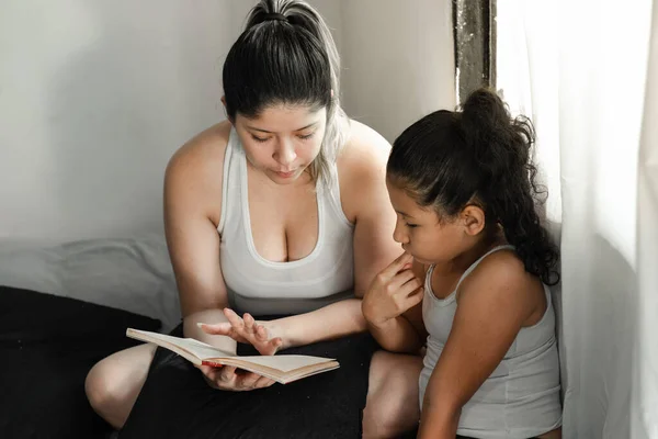 Latina Leyendo Libro Pequeña Hija Morena Mientras Mira Atentamente Con — Foto de Stock