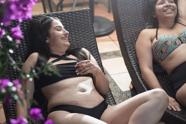 2人の若いラテン系の女性がプールラウンジャーでリラックスし チャットと笑いを楽しんでいます — ストック写真