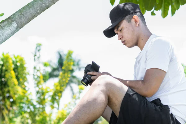 Ağacın altındaki çimlerin üzerinde oturan Latin etkileyicisi kamerasıyla çektiği fotoğraflara bakıyor.