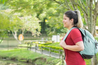 Güzel, genç bir Latin üniversiteli kadın, büyük bir çanta taşıyor, gülümsüyor ve parkta yürüyor.