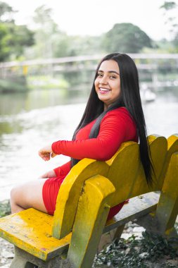 Genç üniversite öğrencisi gölün kenarında sarı bir sandalyede oturuyor, çok mutlu ve gülümsüyor..