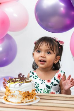 Küçük Latin esmer kız doğum günü pastasının önünde mutlu bir şekilde gülümsüyor, etrafında balonlar var.