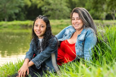 Anne ve kızı gölün yanındaki çimenlikte otururken gülümsüyorlar.