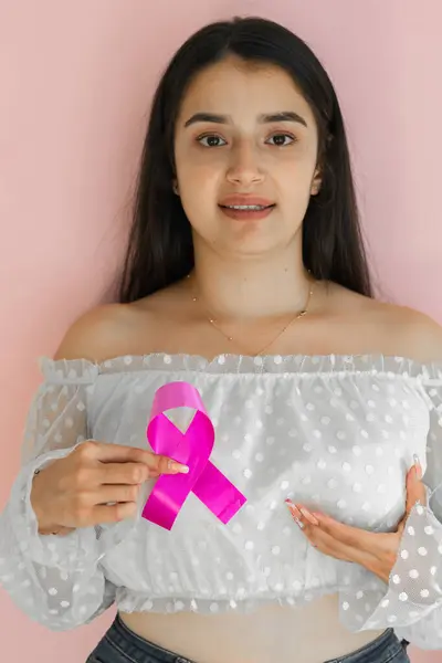 latina woman holding a pink ribbon with one hand while clutching her breast with the other hand. fight against cancer