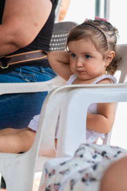 Küçük Latin kız, beyaz bir sandalyede oturmuş yemek beklerken kafasını kaşıyordu.