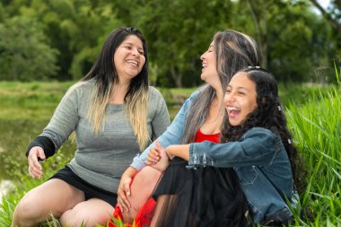 Latin arkadaşlar çimenlerde oturmuş kahkahalarla gülüyorlar ve birbirleriyle takılıyorlar.