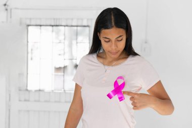 young Latin brunette woman looking and pointing with her finger at the pink ribbon that she has stuck to her chest, a symbol of the fight against breast cancer