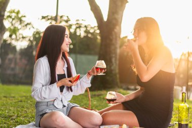 İki Latin arkadaş konuşuyor, gülüyor, meyve yiyor, şarap içiyor, parkta oturuyor, gün batımında..