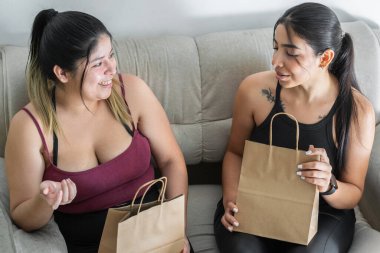 Genç Latin kadınlar ellerinde kahverengi torbalarla alışveriş yaptıktan sonra koltukta oturuyorlar.