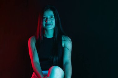 Uzun saçlı, uzun boylu, kırmızı ve mavi renkli stüdyo ışığı olan güzel, genç bir Latin kadın, kurnazca gülümsüyor ve utangaç bakışlarıyla başka yere bakıyor..
