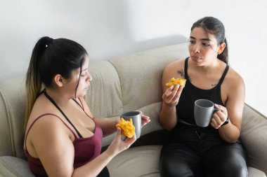 Genç Latin bir kadın kahve içip Kolombiya usulü etli börek yerken arkadaşına bakıyor.