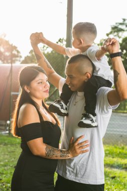 Parkta çocuğuyla yürüyen genç Latin bir çift. Kadın kocasına bakarken erkek çocuğu omuzlarında taşıyor. Gün batımında arka planda.