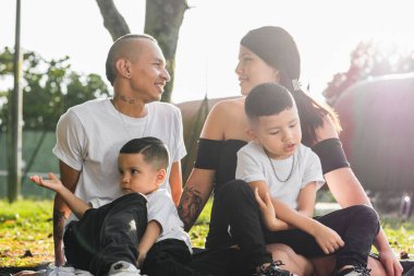 Parkta kucağında iki çocuğu olan genç Latin ebeveynler birbirlerinin gözlerinin içine bakıp gülümserken dikkat dağıtıcı bir şekilde oynuyorlar.