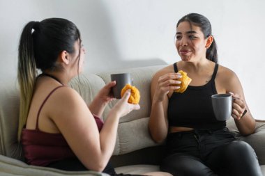 İki genç Latin arkadaş kanepede oturup kahve içerken sohbet ediyor ve abur cuburlarını yiyorlar. Kolombiya usulü etli turta yiyorlar.