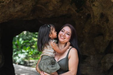 Gülümseyen anne, genç Latin kızını gölgeli bir hayvanat bahçesi mağarasında öpüyor.