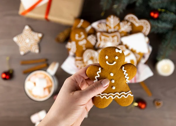 A female hand holds a gingerbread man on a background of a gingerbread basket, a cup of cocoa with marshmallows, a gift box and Christmas tree branches.