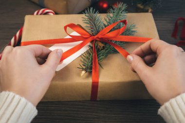 Noel ağacı dallarıyla süslenmiş bir hediye kutusuna kırmızı kurdele bağlayan eller ve arka planda tarçın çubuğu. Boşluğu kopyala