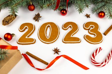 Beyaz arka planda 2023 numara. Yakınlarda Noel ağacı dalları, kırmızı toplar, koniler, kurdeleler, bir lolipop, tarçın ve yıldız anason var.