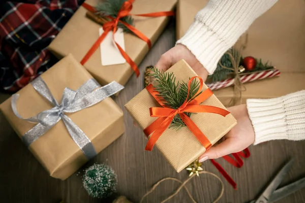 Noel süslemesinin arka planında kırmızı kurdeleli ve Noel ağacı dallı hediye kutusu olan kadın eller. Tatil için hediye hazırlama konsepti.