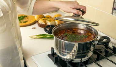 Bir kadın aşçı mutfak sobasında bir demlik çorbanın kapağını açar.