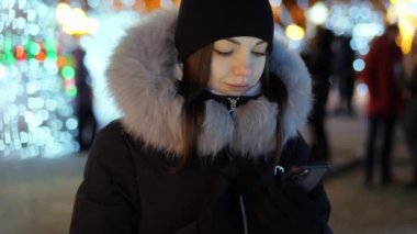 Kadın telefonu merkez Noel ağacının arka planında kullanıyor, arka planda ışıklar ve çelenkler, baskılar, tebrikler ve sosyal ağlar.