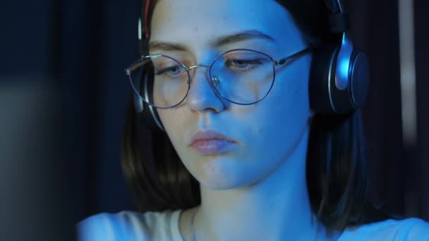 メガネとワイヤレスヘッドフォンを持つ十代の女の子のクローズアップ肖像画は モニターを見て 夜遅くにゲームをプレイ — ストック動画
