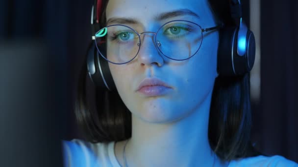 メガネとワイヤレスヘッドフォンを持つ十代の女の子のクローズアップ肖像画は モニターを見て 夜遅くにゲームをプレイ — ストック動画