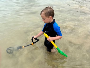 Bir çocuk plajda hazine arıyor, metal dedektörüyle.