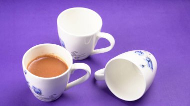 Porselen bir fincan lezzetli sıcak içecek ve mor arka planda boş bardaklar.          