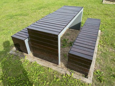 Parkta bankları olan ahşap bir piknik masası. Masa bir piknik için hazırlandı, kırmızı kareli masa örtüsü, tabaklar, bardaklar ve yiyecek. Masada oturan, konuşan ve gülen iki kişi var. Parktaki ağaçlar yeşil ve yemyeşil.